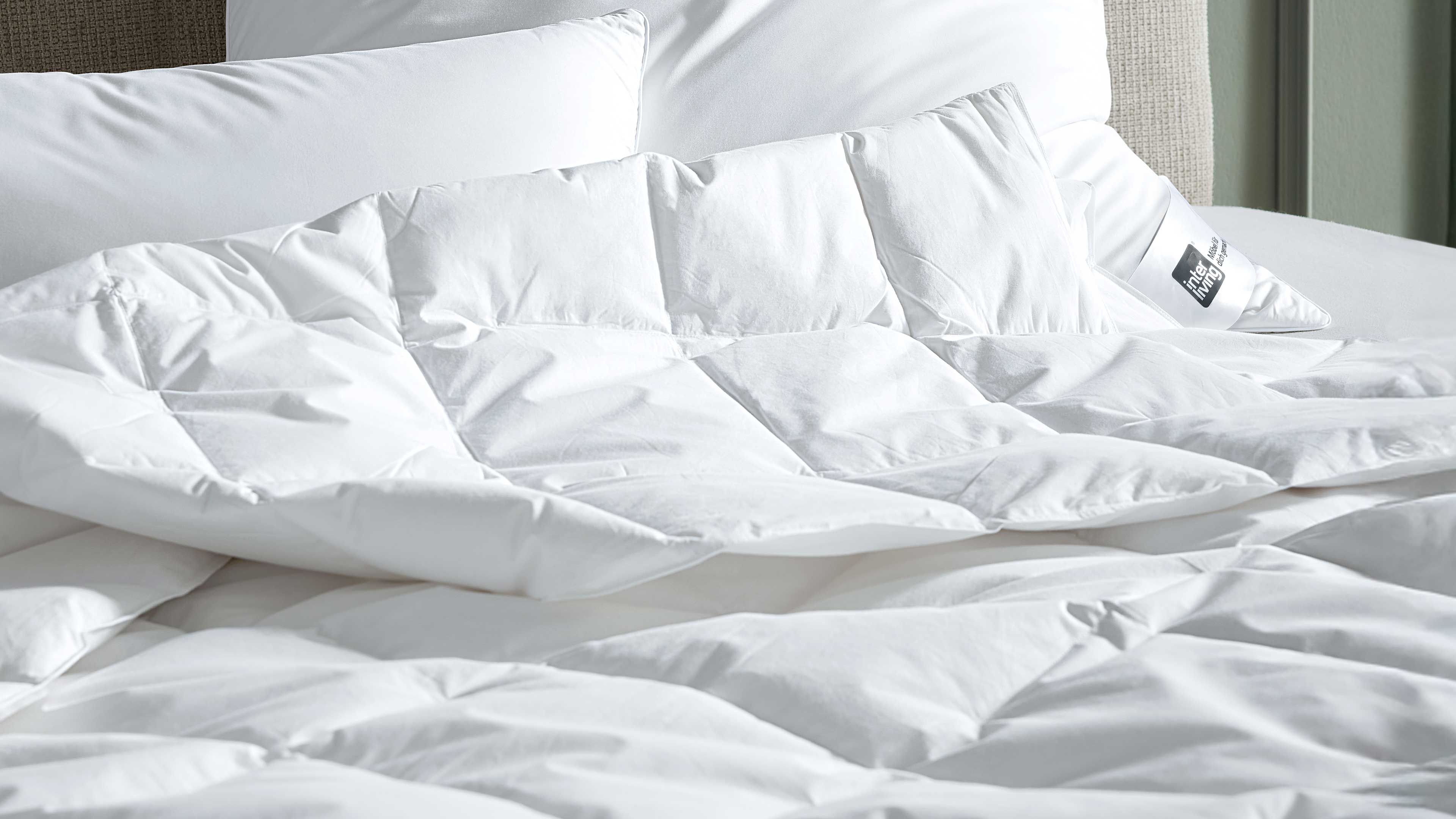 Gesteppte weiße Bettdecke liegt aufgefaltet auf Matratze, mehrere weiße Kissen am Kopfende, eines zeigt ein sichtbares Etikett.