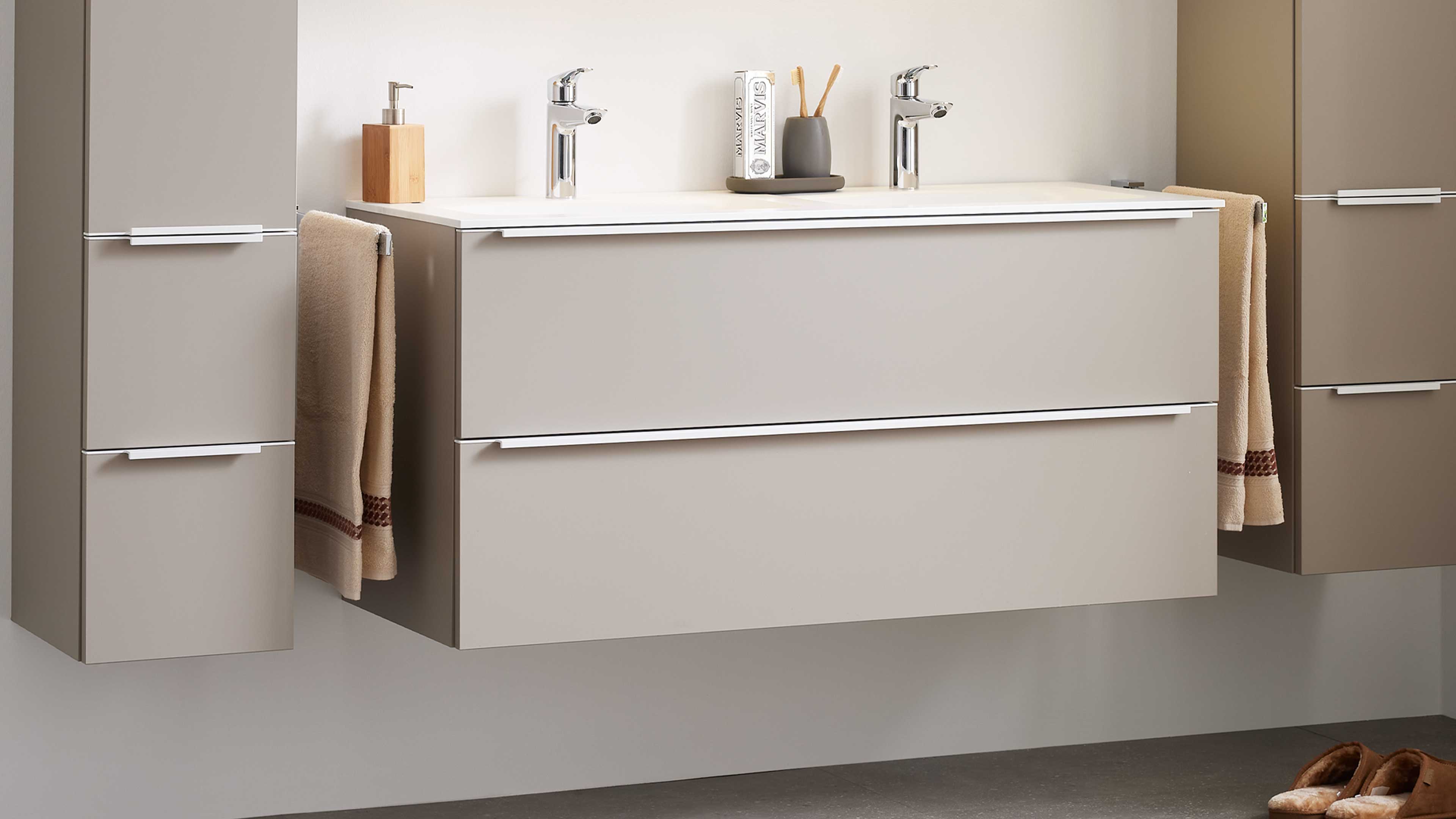 Wandhängender beige Doppelwaschtischunterschrank mit zwei verchromten Armaturen, zwei Schubladen, seitlichen Hochschränken und Handtüchern.