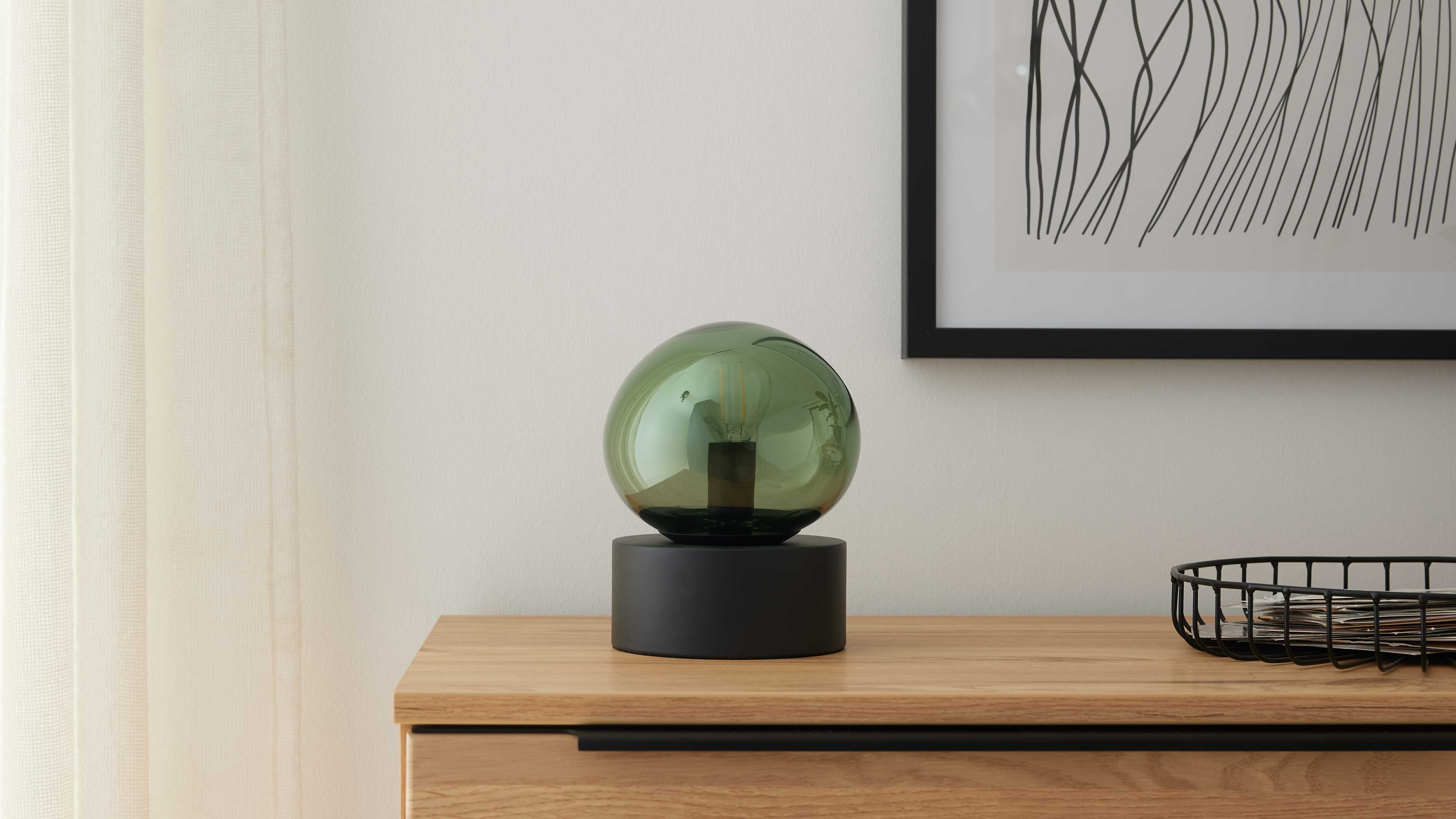Grüne Glaskugel-Lampe auf schwarzem Sockel steht auf Holzkommode; rechts Drahtkorb mit Papieren, links helle Gardine.