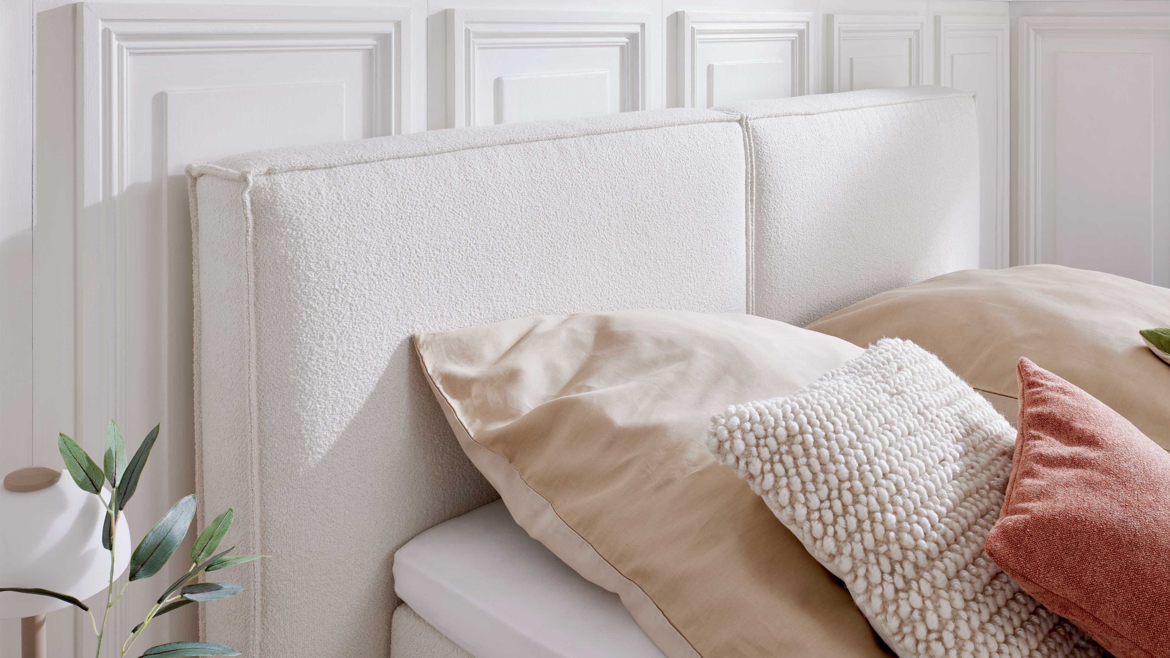 Zweiteiliges weißes Polsterkopfteil vor vertäfelter Wand mit beiger Bettwäsche, einem creme-texturierten Zierkissen und rostfarbenem Kissen.