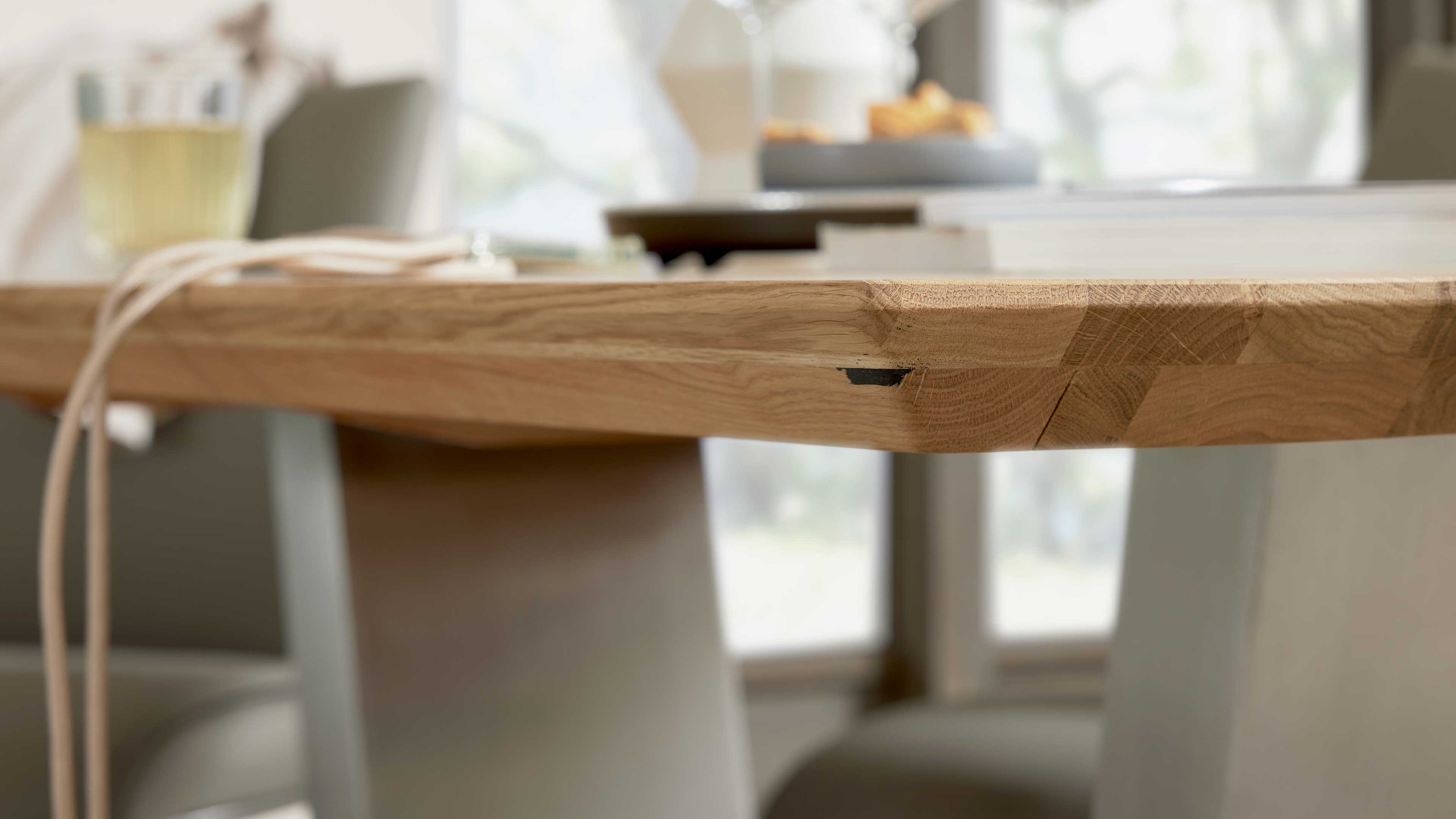 Helle Holz-Tischplatte mit sichtbarer Kante und dunkler Markierung, im Hintergrund unscharfe Stühle, ein Glas und zwei Kabel.