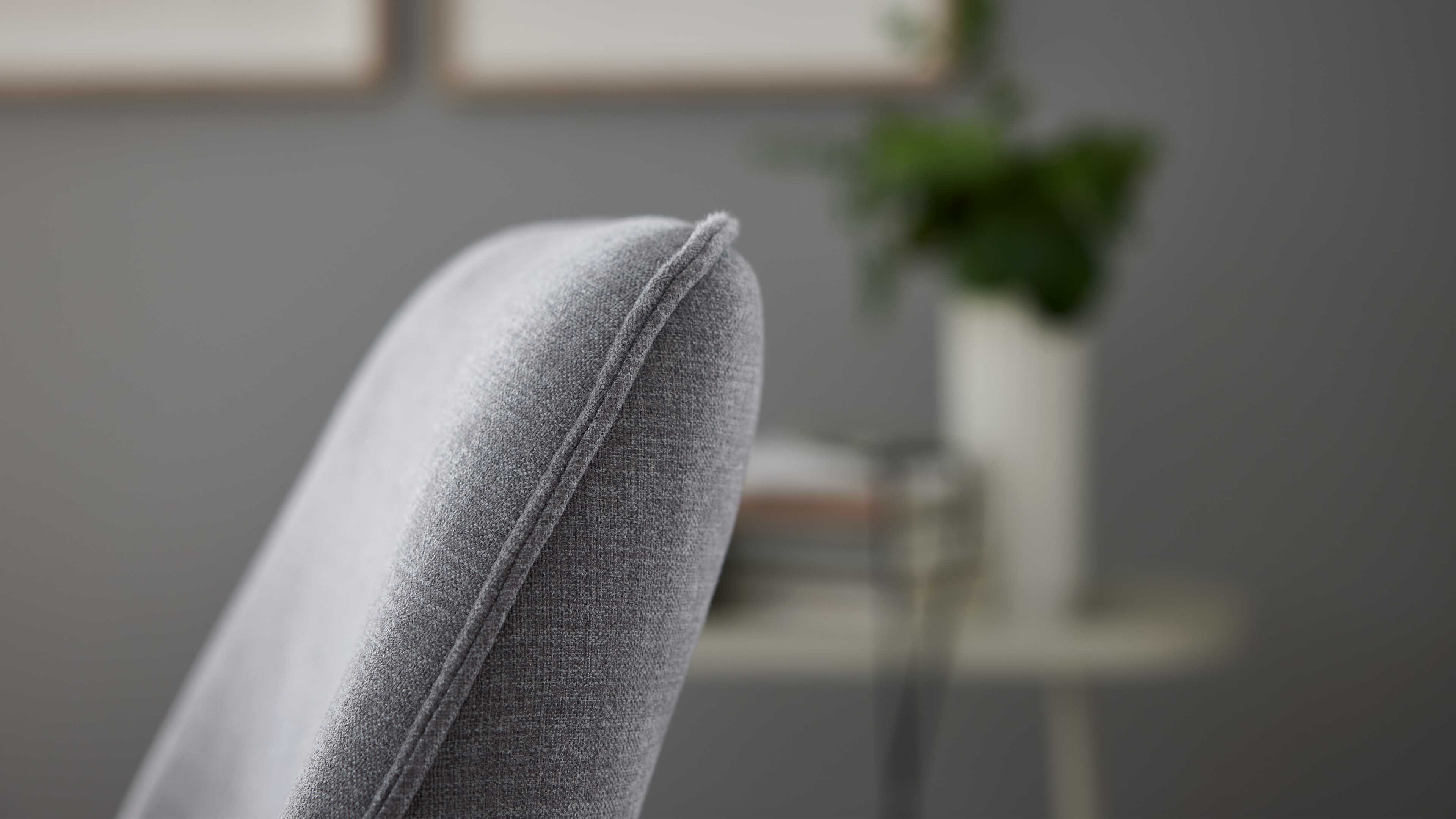 Grau bezogene Rückenlehne eines Stuhls mit sichtbarer Kedernaht, unscharf im Hintergrund Beistelltisch mit Büchern und Vase.